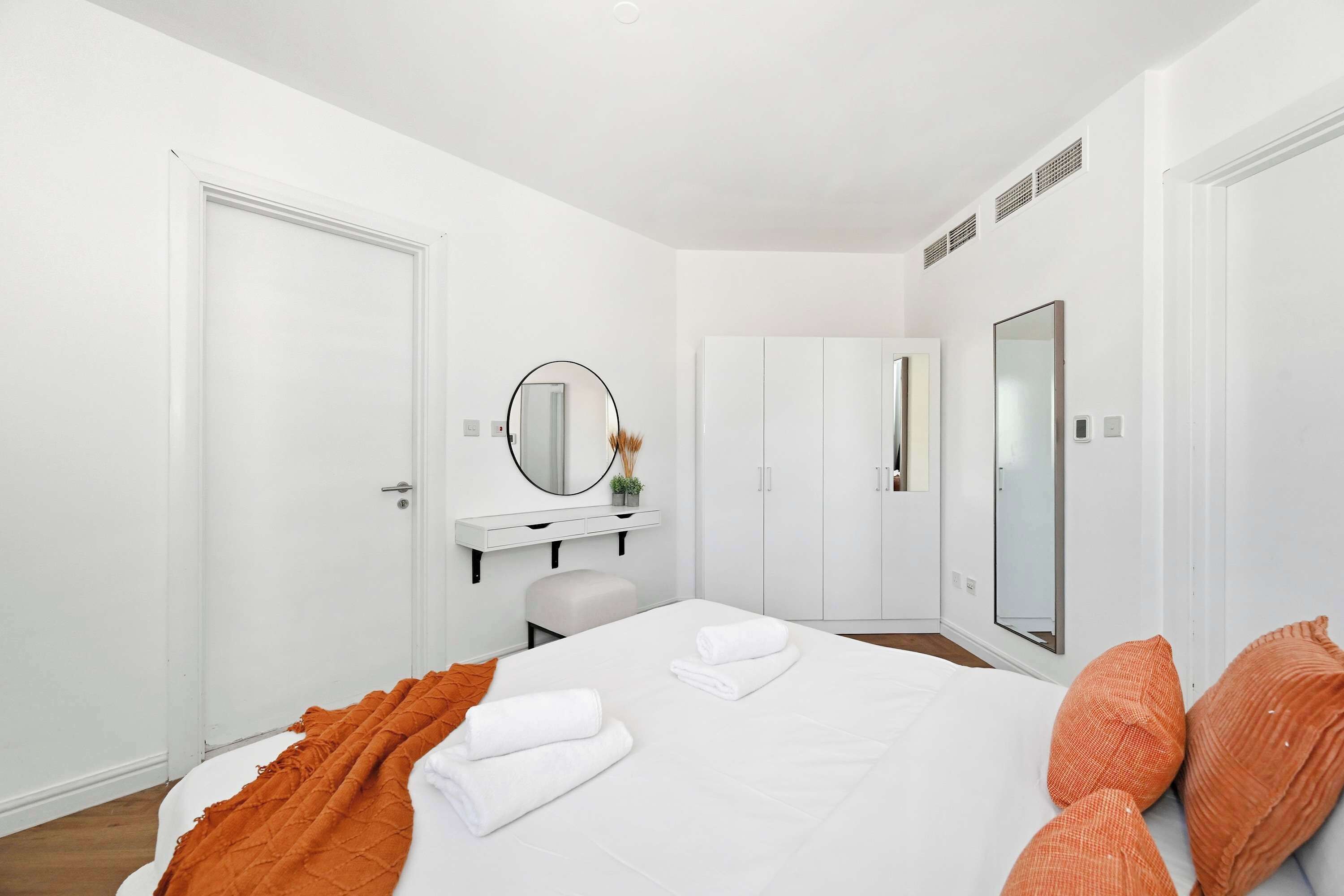 A modern hotel room with a large bed, white walls, a white wardrobe, a vanity with a mirror, and a small seating area.