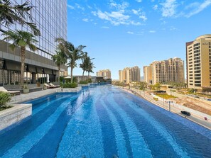 A swimming pool with lounge areas, palm trees, and modern buildings in the background.