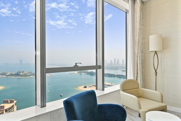 A room with a large window offering a view of a body of water and distant cityscape. There are two chairs and a lamp.