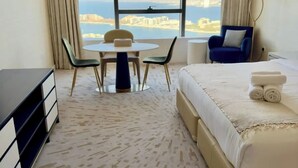 A hotel room with a bed, a desk, a chair, and a view of a cityscape through large windows. - Gorgeous luxury studio - Exquisite Palm View (Palm Jumeirah)
