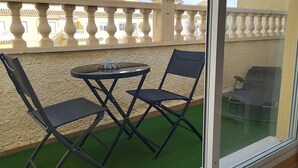 A balcony with a glass door, two folding chairs, a small round table, and a view of buildings in the background. - Bungalow with pool, private patio of 17m2 and terrace (Playa del Carabassi) (Gran Alacant)