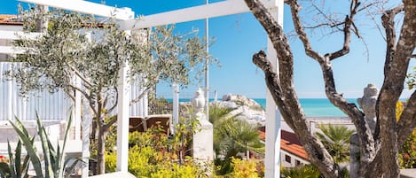 A terrace with white railings, olive trees, and a view of the sea.