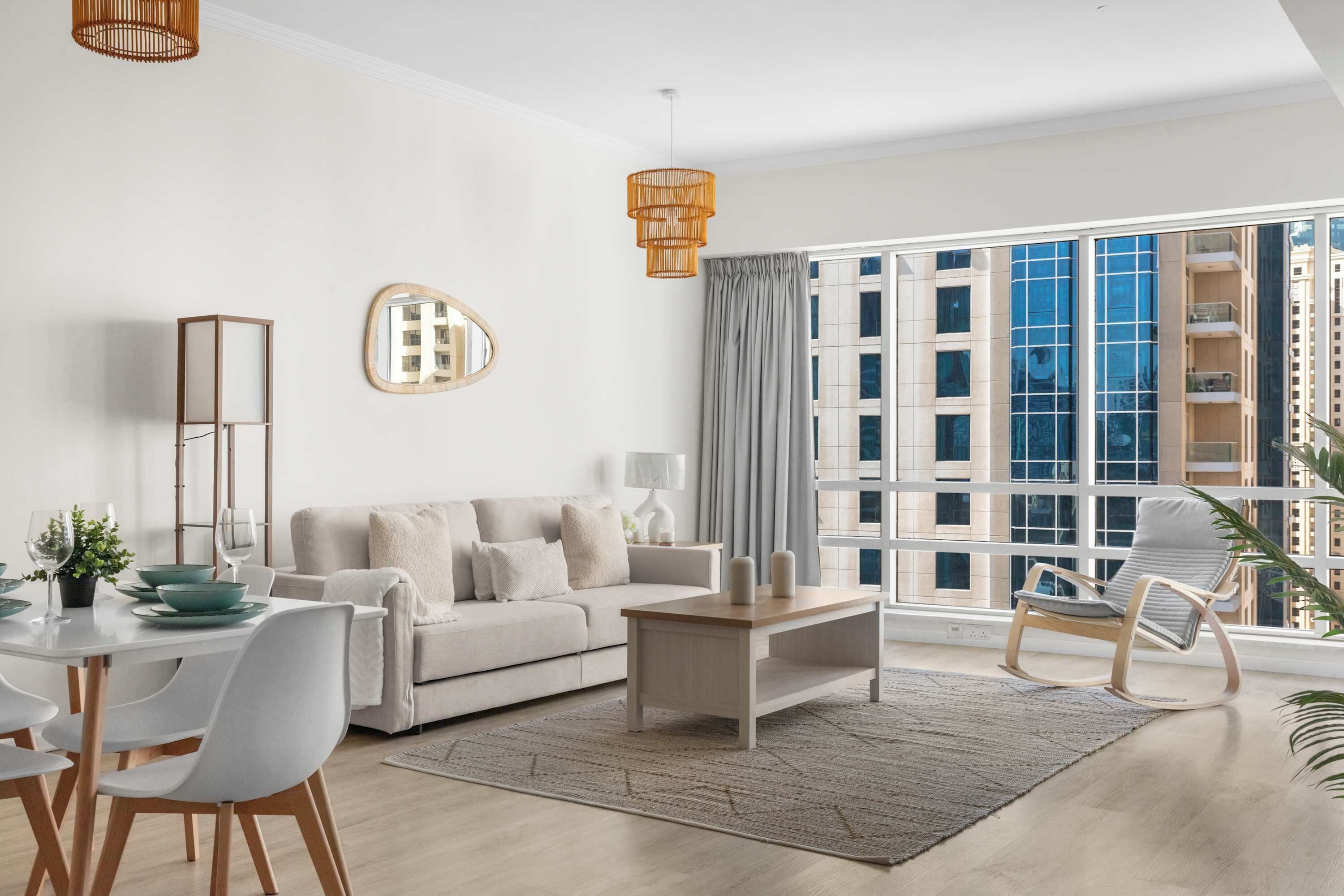 A modern living room with a sofa, coffee table, and chairs, a dining area with a table and chairs, and large windows with a view of buildings.