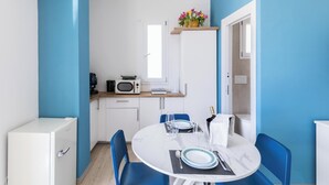 Cocina compacta con armarios blancos, microondas y cafetera. Cuenta con un comedor con mesa redonda, sillas azules y refrigerador blanco.