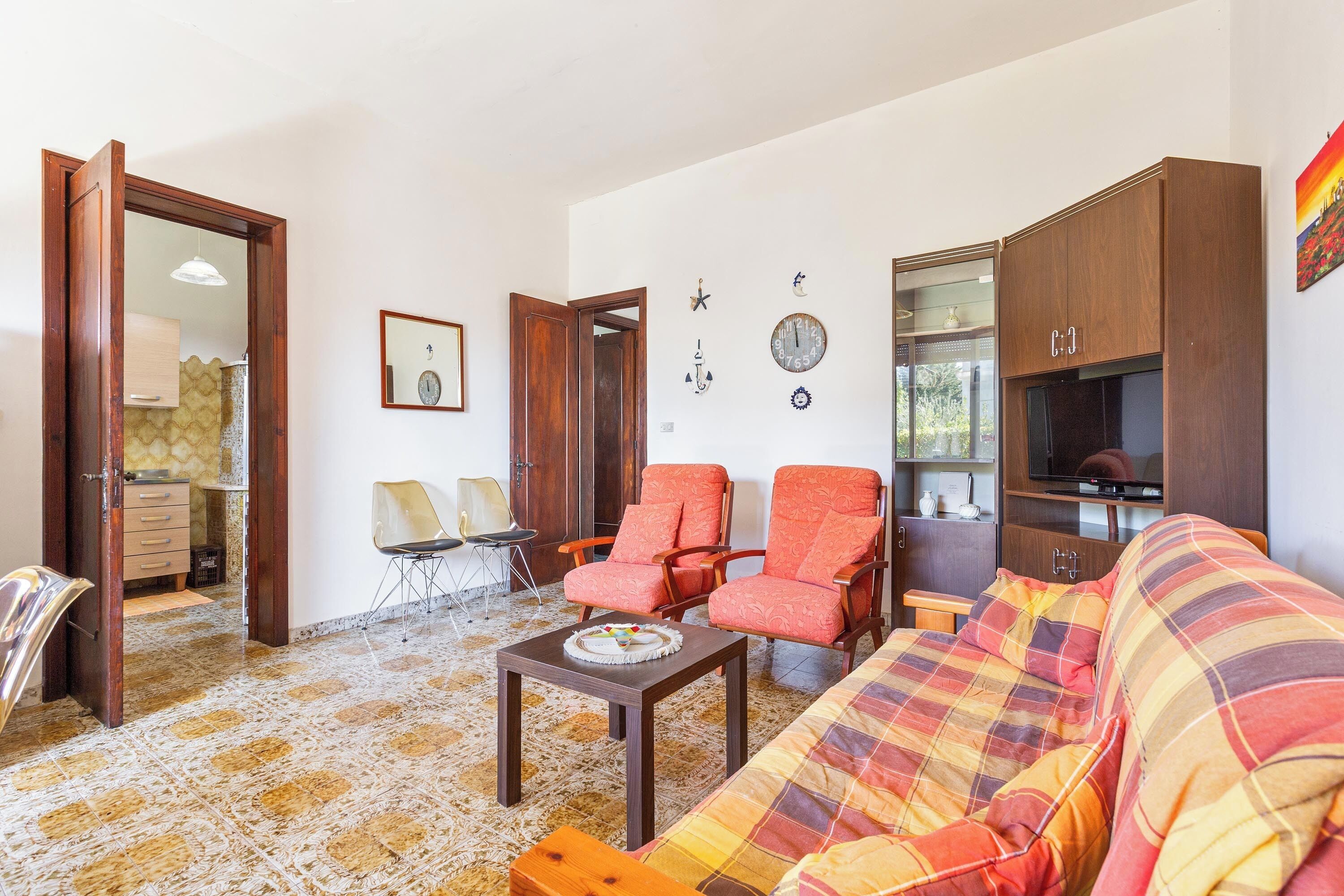 Un salon comprenant un canapé, deux fauteuils, une table basse et un téléviseur. Une porte donne accès à une autre pièce, et il y a une horloge murale.