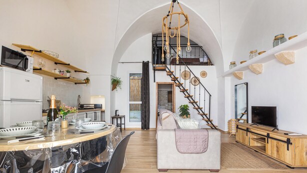 A spacious living area with a high ceiling, a wooden staircase, a sofa, a TV stand, and a kitchen area with a microwave and shelves.
