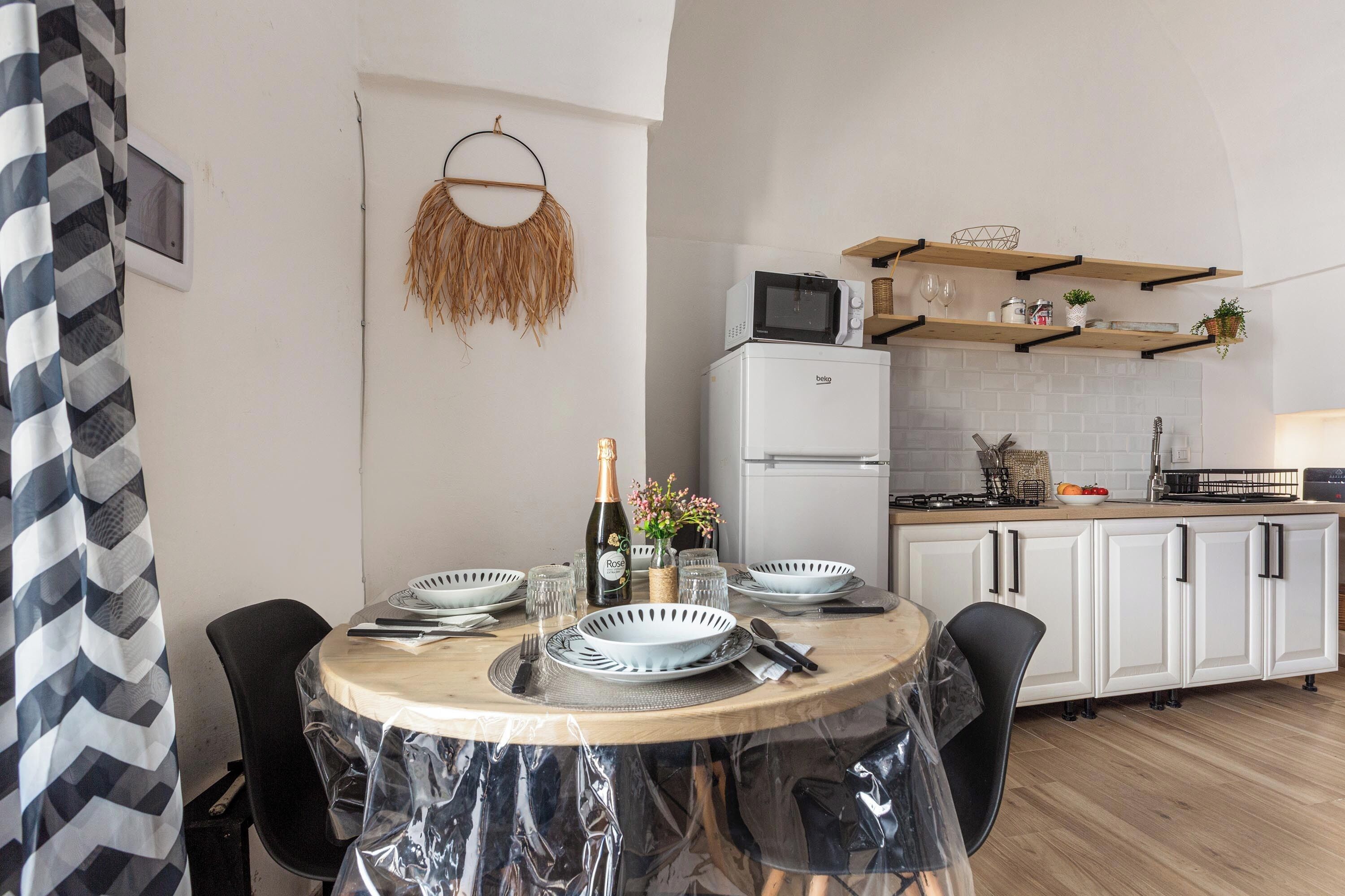 Una cocina moderna con armarios blancos, un microondas y un horno empotrado. Hay una mesa de comedor con sillas y un mantel de plástico transparente.