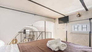 A bedroom with a brown quilted bedspread, a rolled white towel on the bed, a TV mounted on the wall, and a window with blinds.