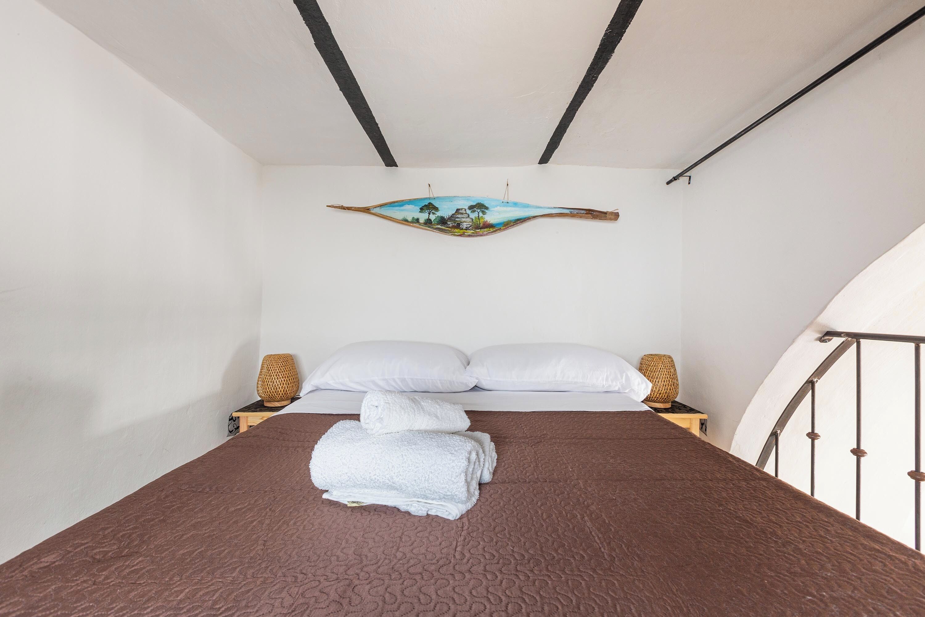 Una cama bien hecha con una colcha marrón texturizada, almohadas blancas y una toalla blanca doblada encima. Hay dos lámparas de noche con bases tejidas y una obra de arte en la pared que representa una escena tropical.