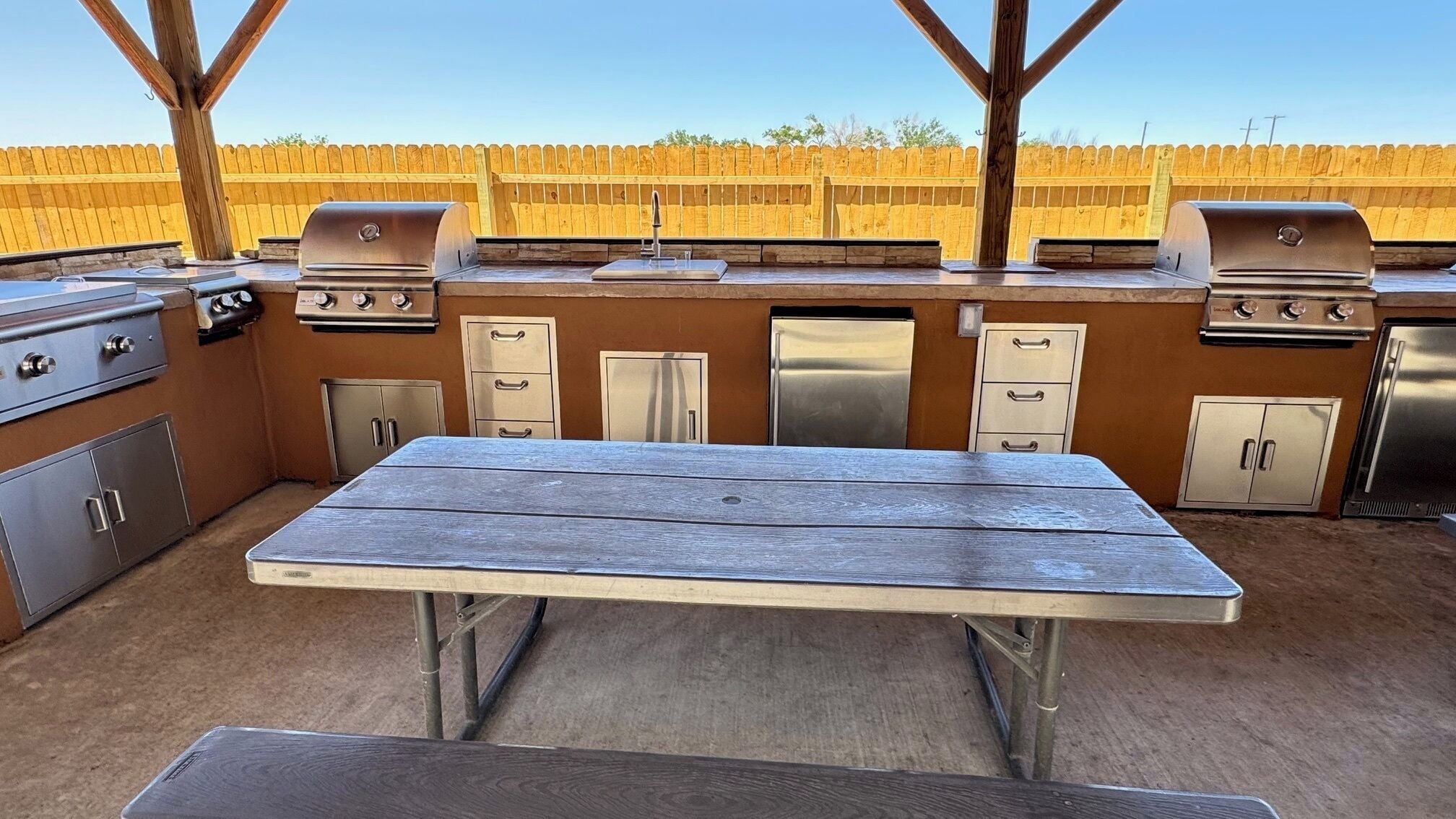 An outdoor kitchen with a wooden table, built-in appliances, and a wooden fence in the background.