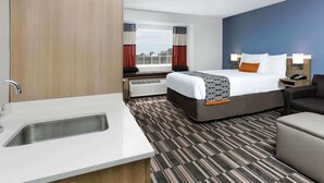 A hotel room with a bed, a sofa, a small table, a window with curtains, a wall-mounted lamp, and a painting. - Permian Studio Apartments (Monahans)