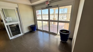 A hallway with a glass door leading outside, two potted plants, and a tiled floor. - Permian Studio Apartments (Monahans)