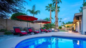 A swimming pool with lounge chairs and umbrellas, surrounded by palm trees and a brick wall. - Private Pool & Putting Green | Fire Pit Patio (Scottsdale)