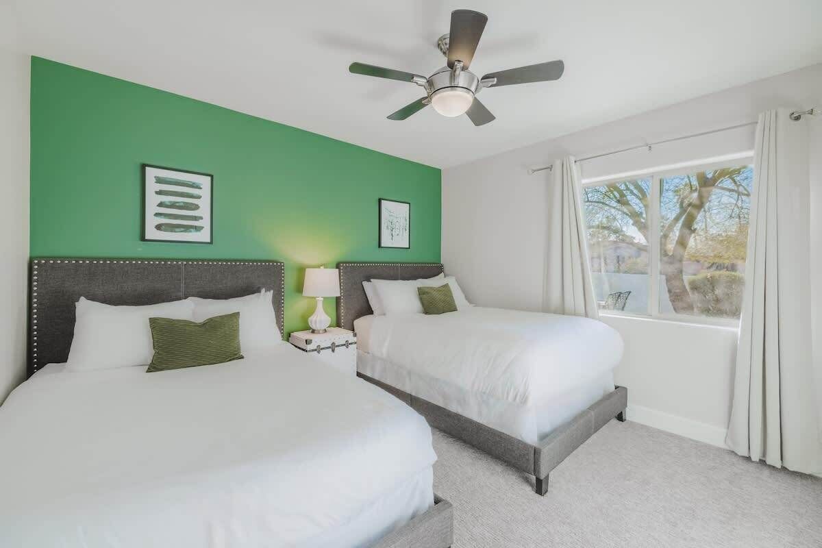 A hotel room with two beds, a ceiling fan, a window with curtains, and artwork on the walls.