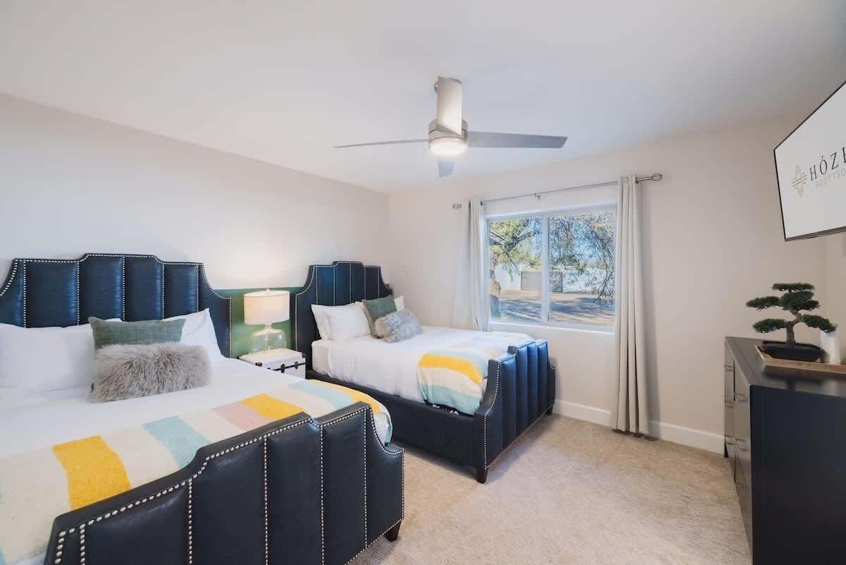 A bedroom with two beds, a ceiling fan, a window with curtains, and a small desk with a potted plant.
