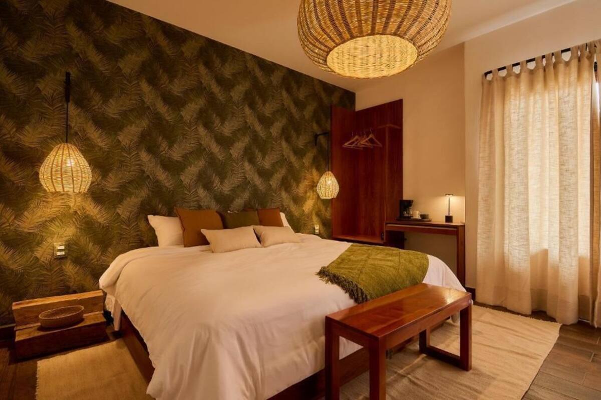 Un dormitorio con una cama, dos mesitas de noche, un banco de madera, una lámpara de fibra natural, una pared con diseño de hojas y una ventana con cortinas.