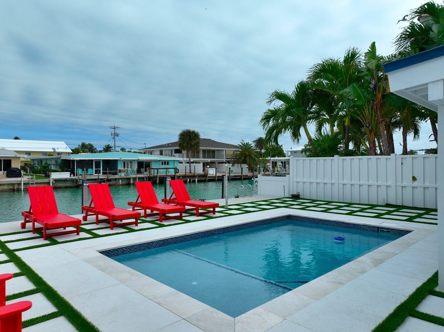 A rectangular pool with red lounge chairs and a white fence.