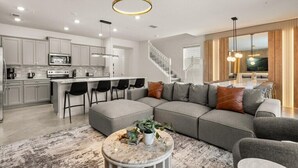 A modern kitchen with a central island, a dining area with a high table, and a living room with a large sectional sofa and a coffee table. - 5 BD/ 4.5 BA/ Windsor Cay (16053 BW) (Clermont)