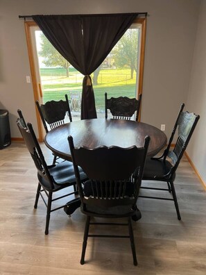 A dining area with a round table and six chairs, a window with curtains, and a view of a green landscape.