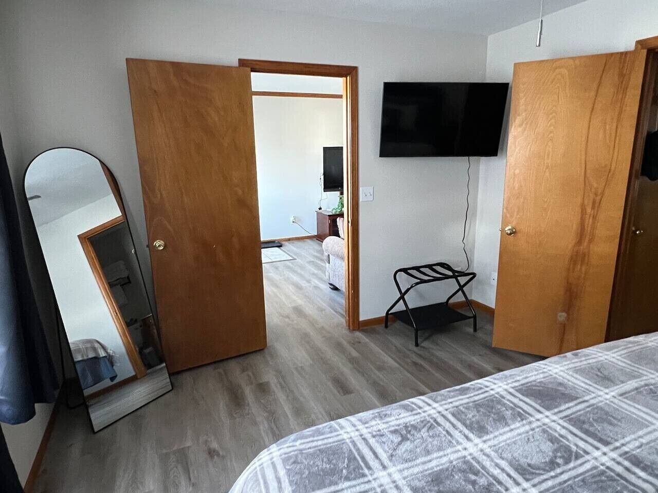 A bedroom with a bed, a wooden wardrobe, a mirror, a television mounted on the wall, and a door leading to another room.