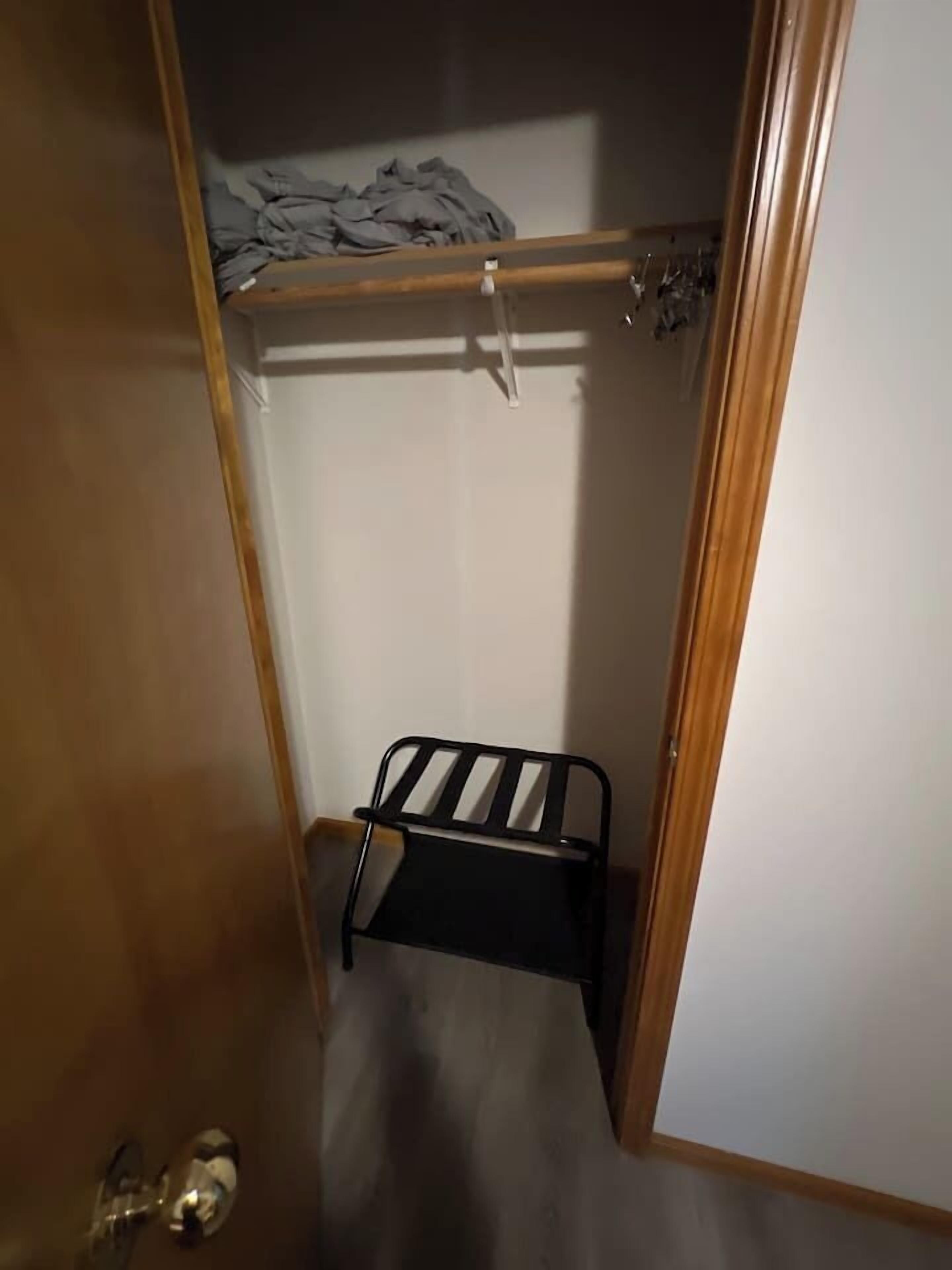 Ein schmales Zimmer mit Holztür, einem kleinen Metallregal und einem Kleiderständer mit gefalteter Kleidung.