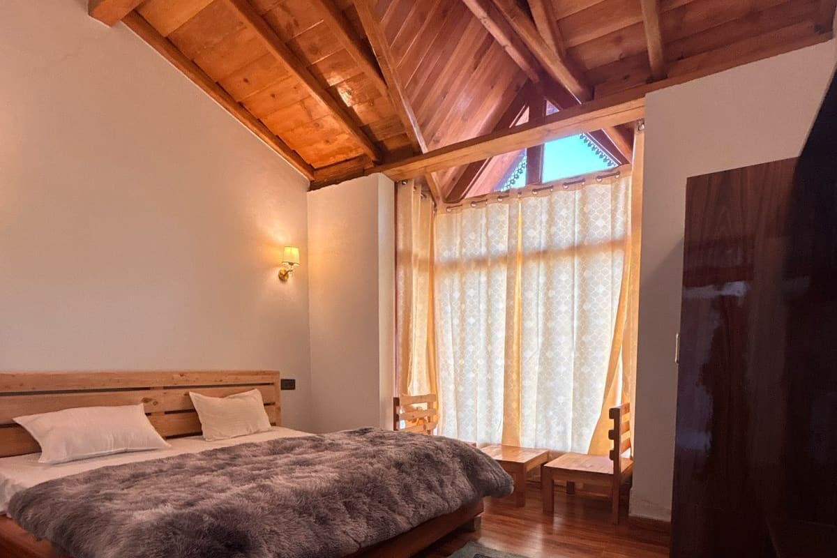 Une chambre avec un plafond en bois, un lit recouvert d’une couverture moelleuse, une table de chevet, une chaise et une fenêtre avec des rideaux.