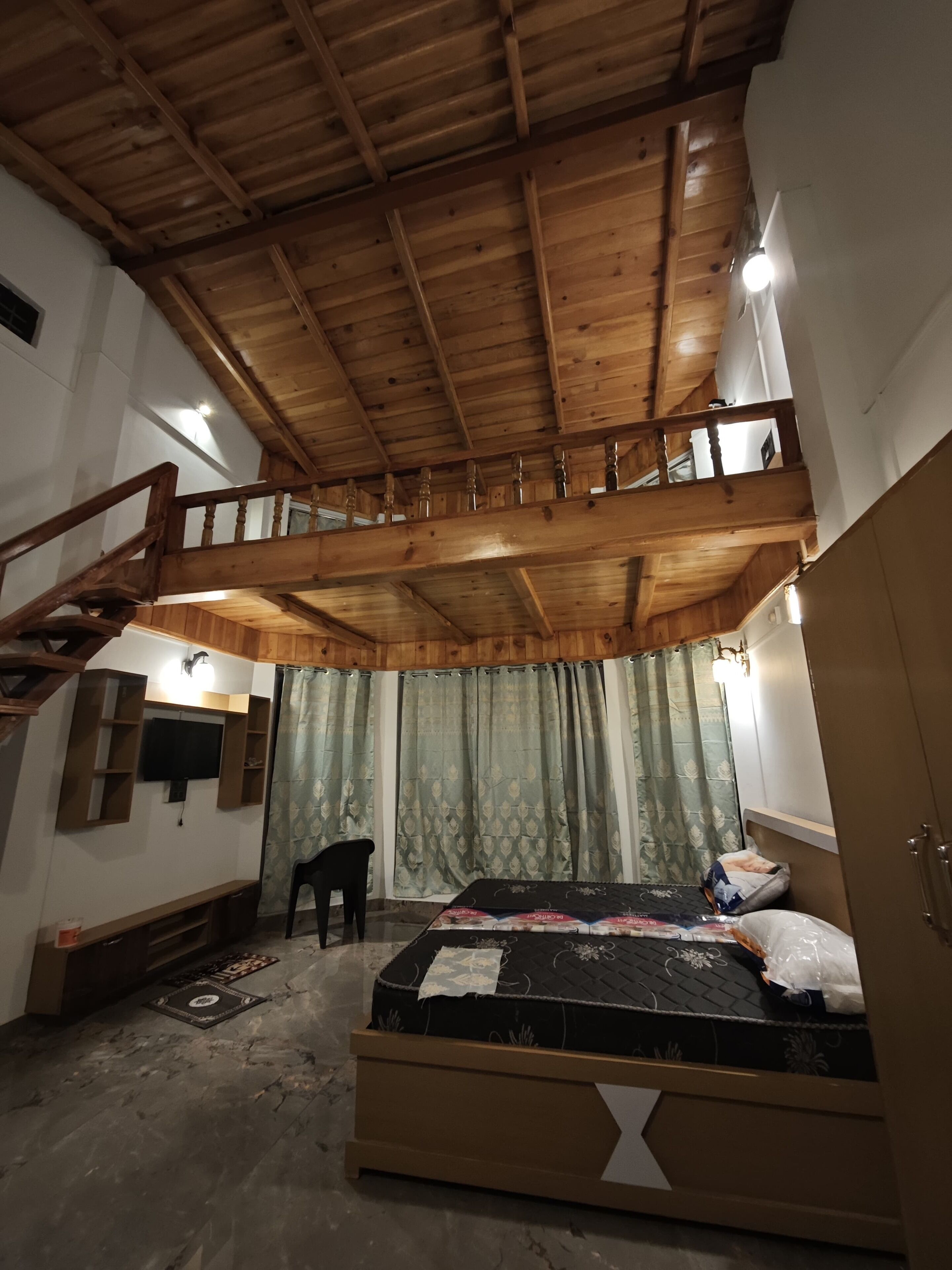 Una habitación con techo de madera elevado, una cama con colchón y almohadas, un televisor montado en la pared y una silla.