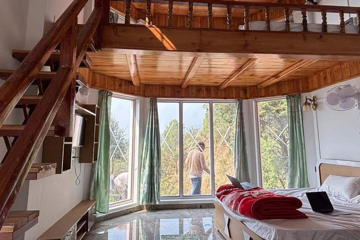 Une pièce avec un escalier en bois, un lit sur lequel se trouve un ordinateur portable, et une vue sur l’extérieur grâce à de grandes fenêtres.
