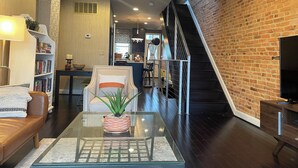 Una sala de estar moderna con una mesa de centro de vidrio, un sofá y una estantería. Se ve una escalera que sube a un nivel superior.