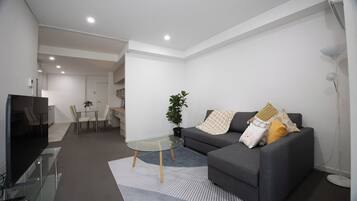 Een moderne woonkamer met een grijze bank, een glazen salontafel, en een eethoek met een witte tafel en stoelen.