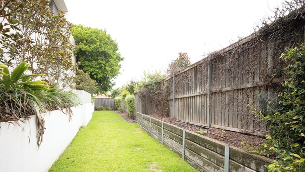 A backyard with a wooden fence, a white wall, and a grassy area. - Kozyguru | Epping | Stylish 1 Bedroom Apartment (Epping)