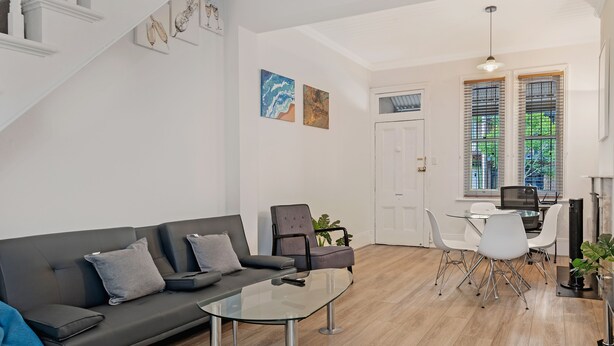 A modern living room with a glass coffee table, grey sofa, and white chairs. There is a staircase with white railings and a door leading to another room. - Kozyguru | Surry Hills | Classic Terraced House (Surry Hills)