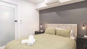 Cama bem arrumada com colcha verde e duas almofadas, abajur e ar-condicionado fixado na parede.