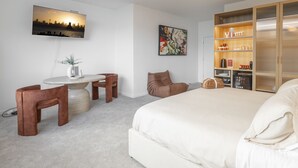 Une chambre d’hôtel moderne comprenant un lit, un bureau avec une chaise, un canapé, une bibliothèque et un tableau représentant un paysage urbain.