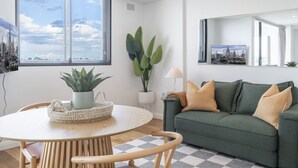 A modern living room with a round wooden table, a green sofa with cushions, and large windows offering a city view. - Kozyguru | Rozelle | Bridge View With Carpark (Rozelle)