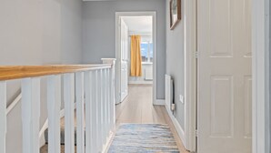 Un couloir doté d’une rambarde en bois, d’un tapis à motifs et d’une porte donnant sur une pièce avec une fenêtre et des rideaux.