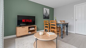 Un salon avec un téléviseur affichant le logo « NETFLIX », une table à manger avec des chaises, une petite table ronde avec des livres et une porte blanche.