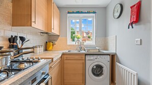 Une cuisine compacte avec des meubles en bois, une cuisinière à gaz, un lave-linge et une fenêtre donnant sur des immeubles.