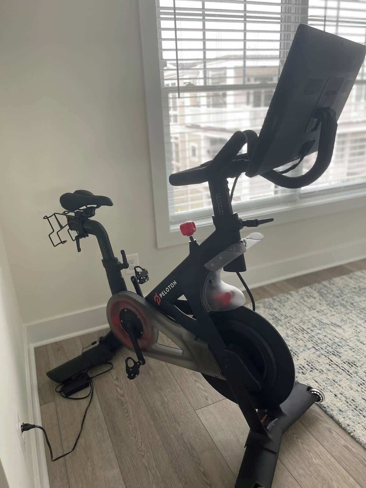 Una bicicleta estática Peloton en una habitación con ventana y suelo de madera.