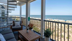 A balcony with a wooden table, cushioned seating, and potted plants. A spiral staircase is visible to the left. The balcony overlooks a beach with people and umbrellas. - Beachfront Luxury Villa in Dewey Beach, Two Master Bedroom (Dewey Beach)