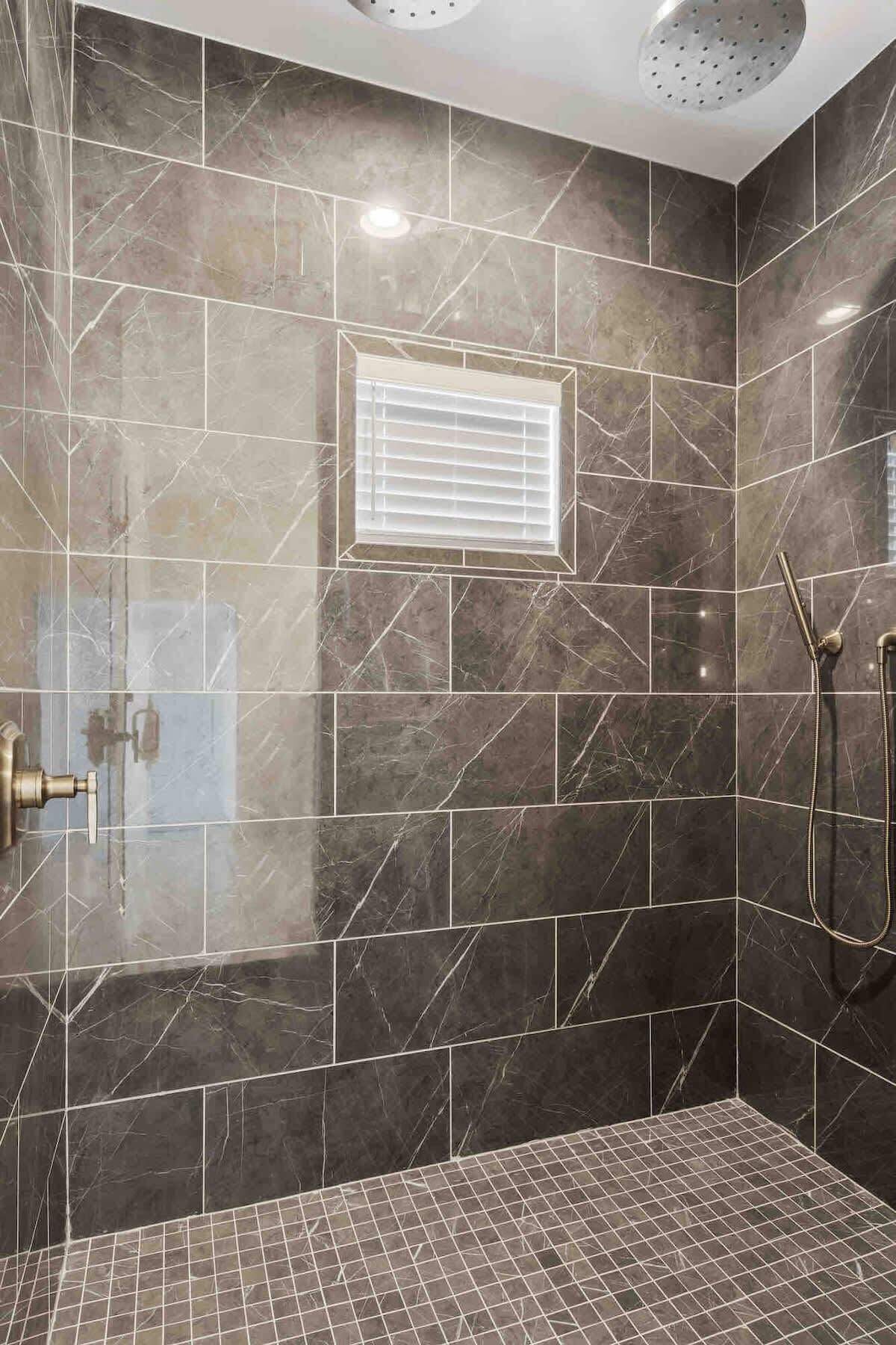 Un espace douche avec des carreaux de marbre foncé, une fenêtre avec des stores et une douchette à main.