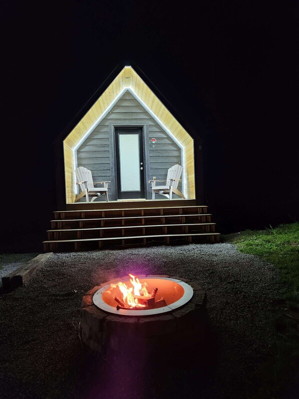 A small cabin with a front door, two chairs on the porch, and a fire pit in front.