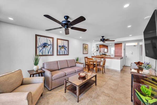 A modern living room with a sofa, coffee table, and ceiling fans. A dining area with a table and chairs is visible in the background.