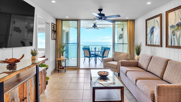 A modern living room with a glass coffee table, a sofa, and a ceiling fan. - Watch Whales from your Bed - Double Master Sunset Suite (Kihei)