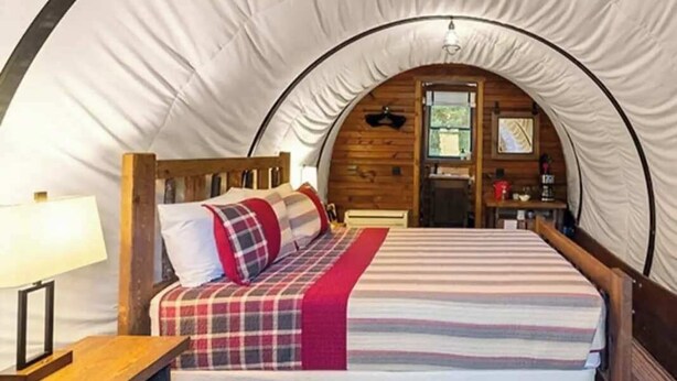 A cozy tented room with a bed, wooden headboard, and a small table with a lamp. - Conestoga Wagon (Wellsboro)
