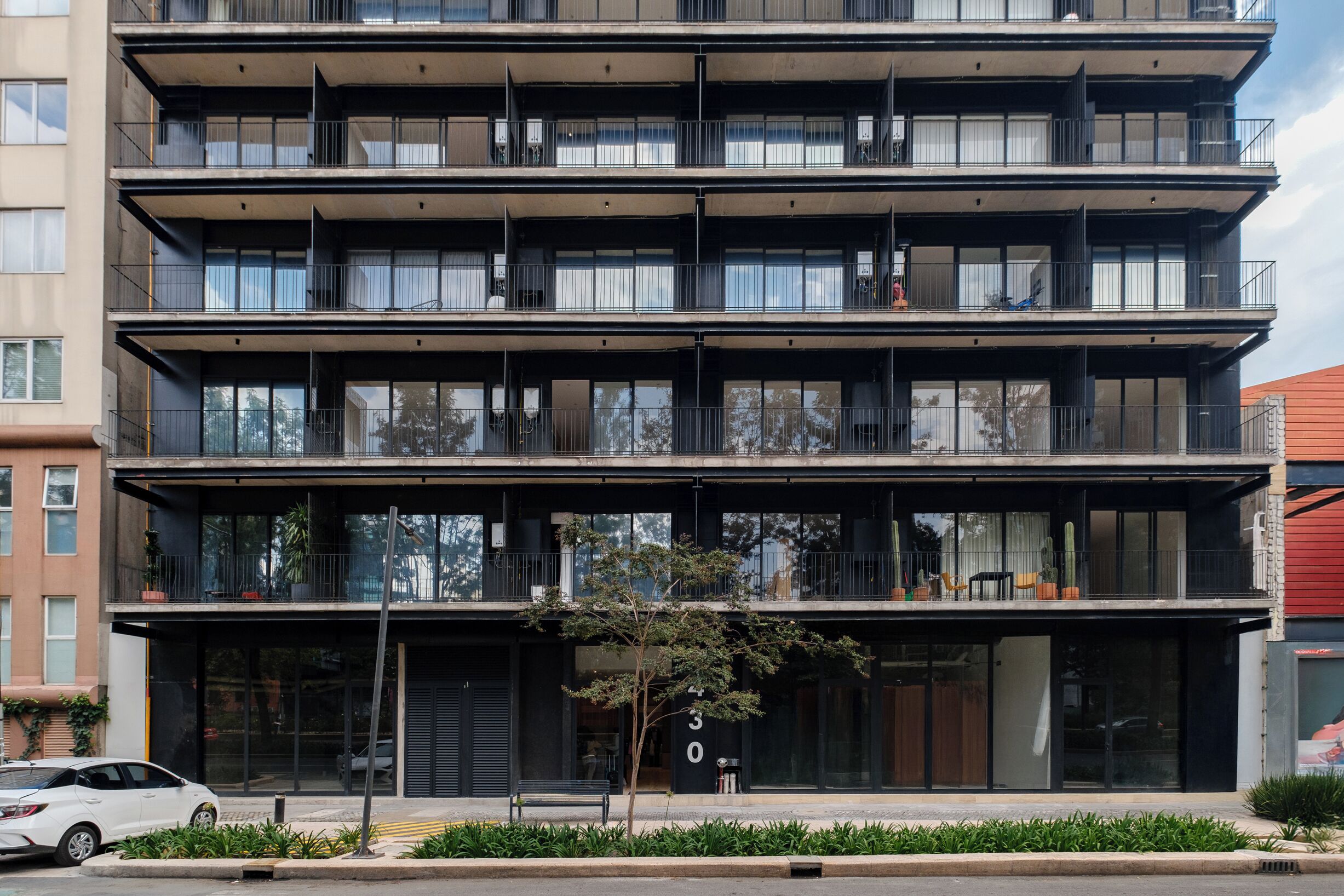 A multi-story building with balconies, large windows, and a small tree in the foreground.