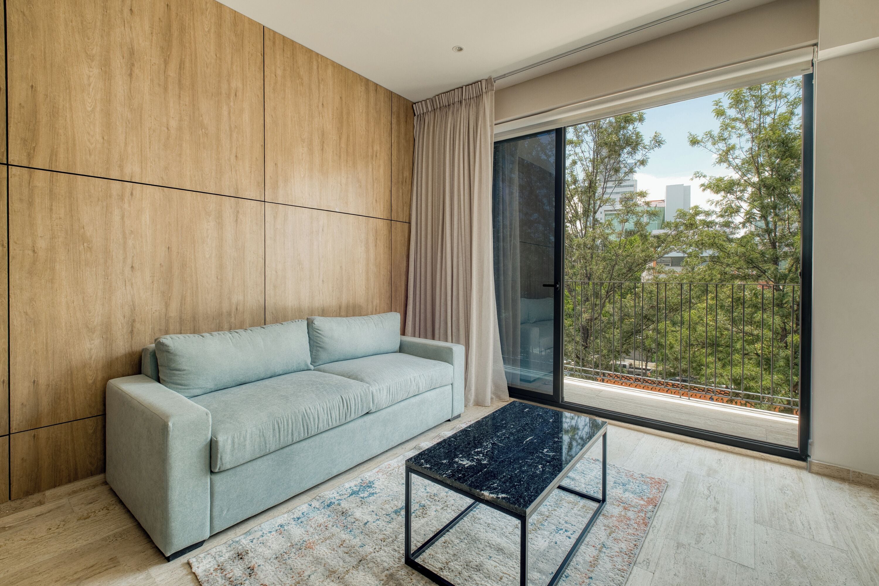 Ein modernes Wohnzimmer mit einem hellblauen Sofa, einem Glastisch und einer großen Schiebeglastür, die zu einem Außenbalkon führt.