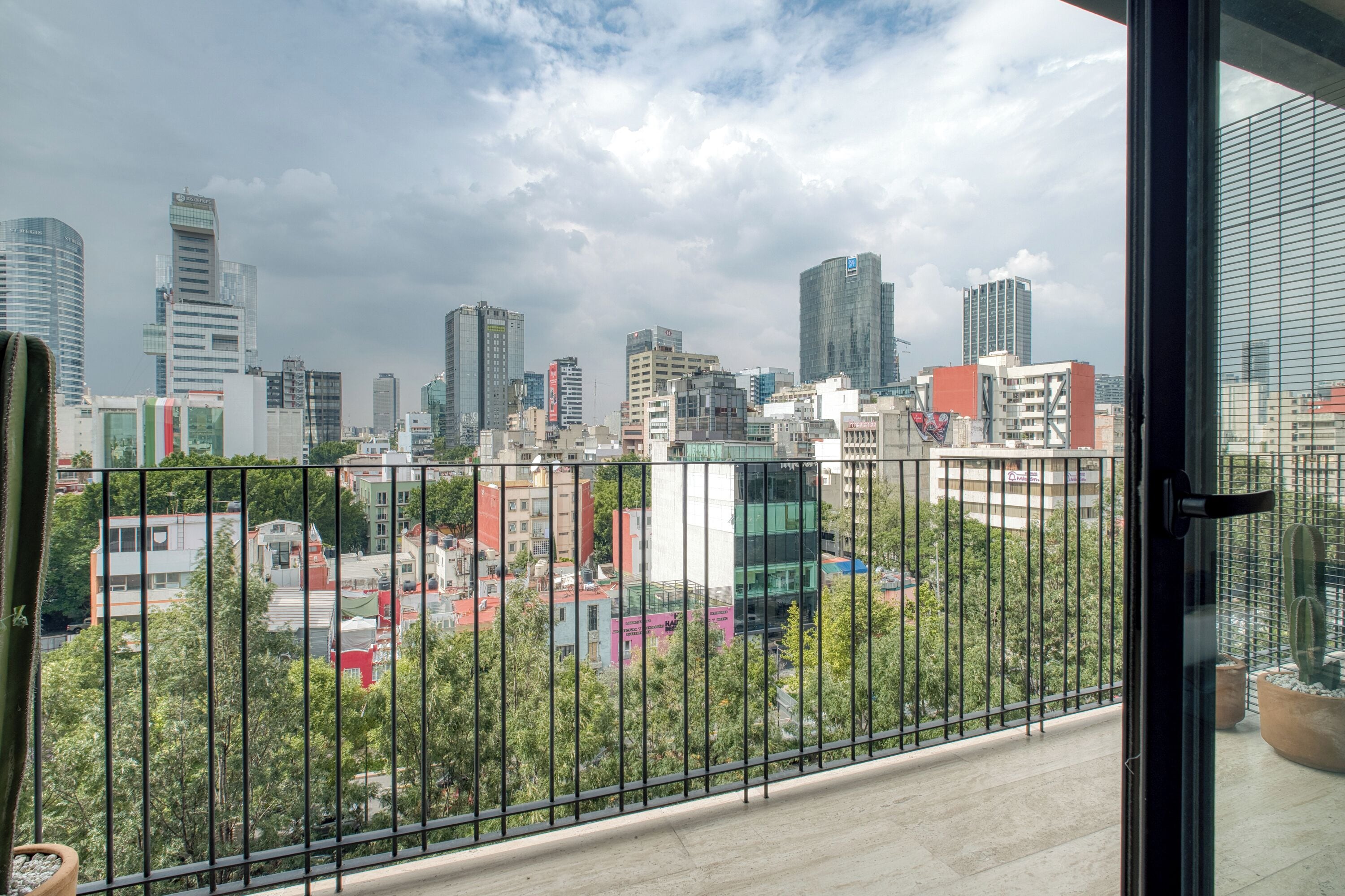 A cityscape view from a balcony with a metal railing, featuring modern buildings and a cloudy sky.