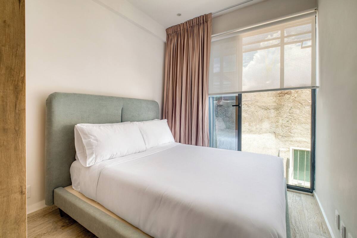 A modern bedroom with a large bed, a window with curtains, and a view of the outdoors.