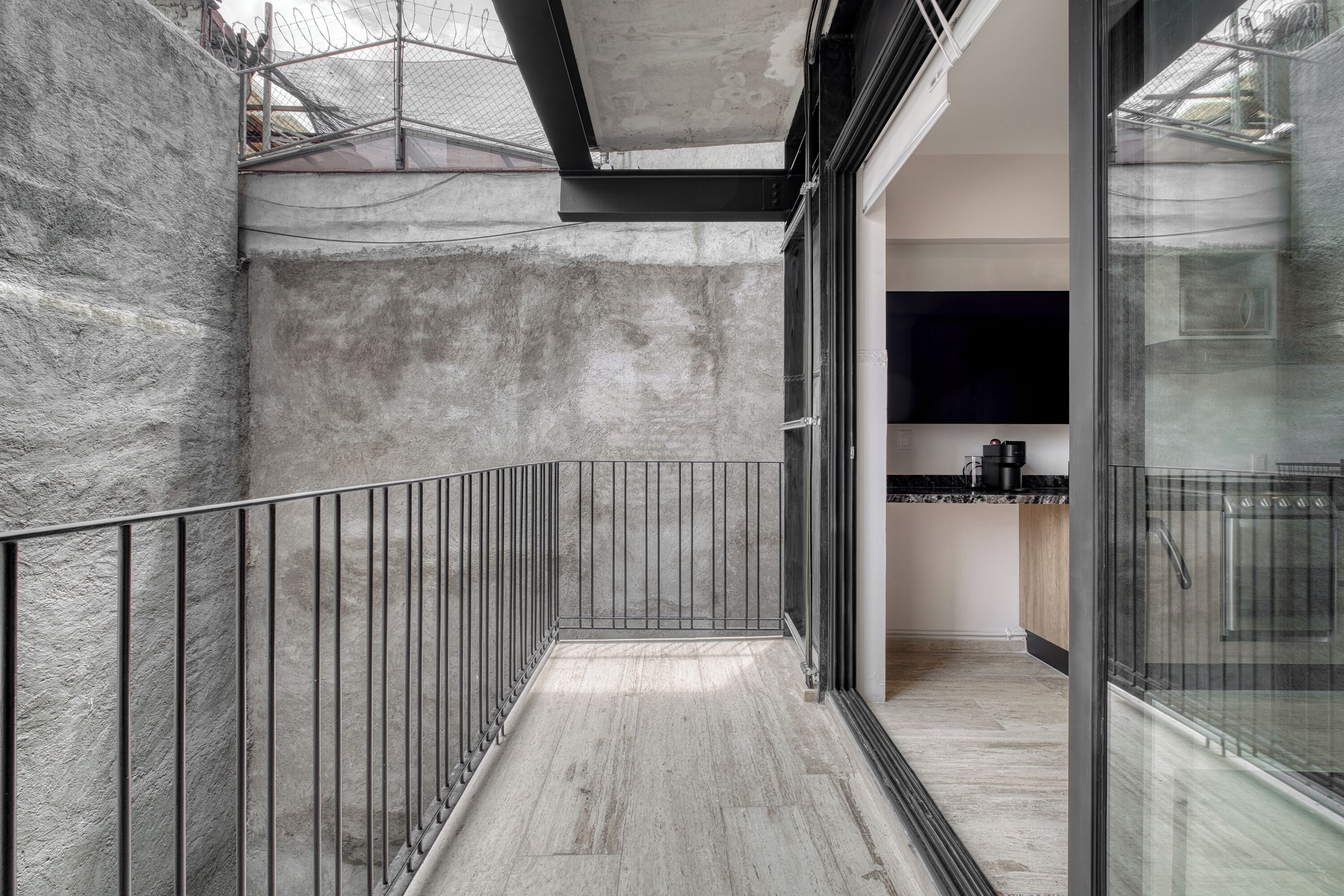 Un balcón pequeño y cerrado con barandilla metálica, pared de concreto y puerta de vidrio que da a una zona de cocina.